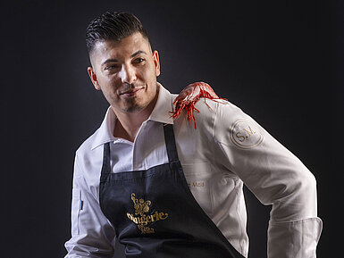 Küchenchef Simone Melis mit Schürze, einer roten Garnele auf der Schulter und einer Zitrone in der Hand.