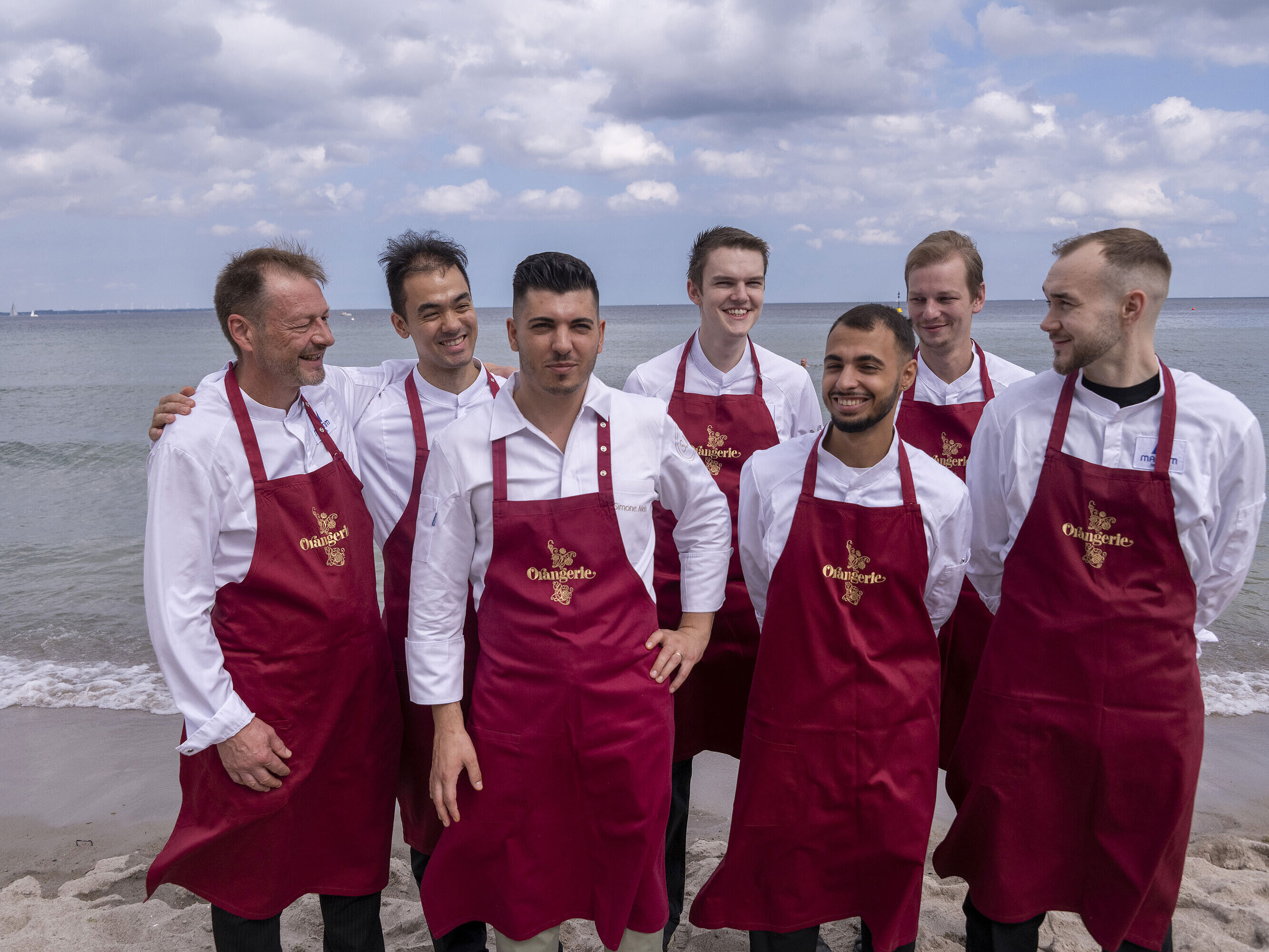 Küchenteam der Orangerie im Maritim Hotel Timmendorfer Strand, am Strand in Kochuniformen und roten Schürzen stehend.