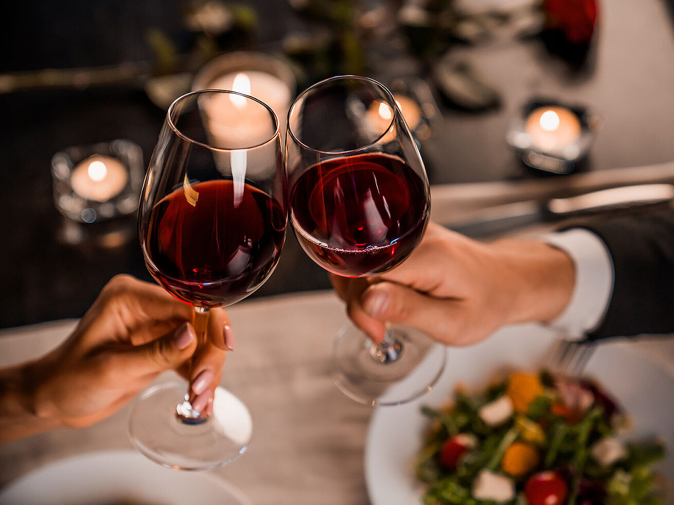 © InsideCreativeHouse - AdobeStock.com Zwei Gläser Rotwein stoßen bei einem romantischen Dinner an