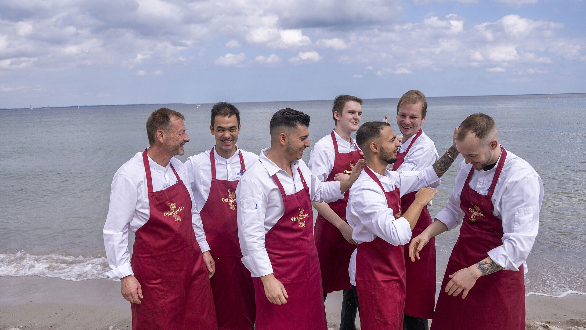 Küchenteam der Orangerie im Maritim Hotel Timmendorfer Strand, am Strand in Kochuniformen und roten Schürzen stehend.