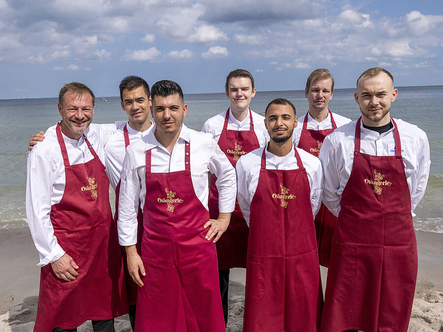 Küchenteam der Orangerie im Maritim Hotel Timmendorfer Strand, am Strand in Kochuniformen und roten Schürzen stehend.