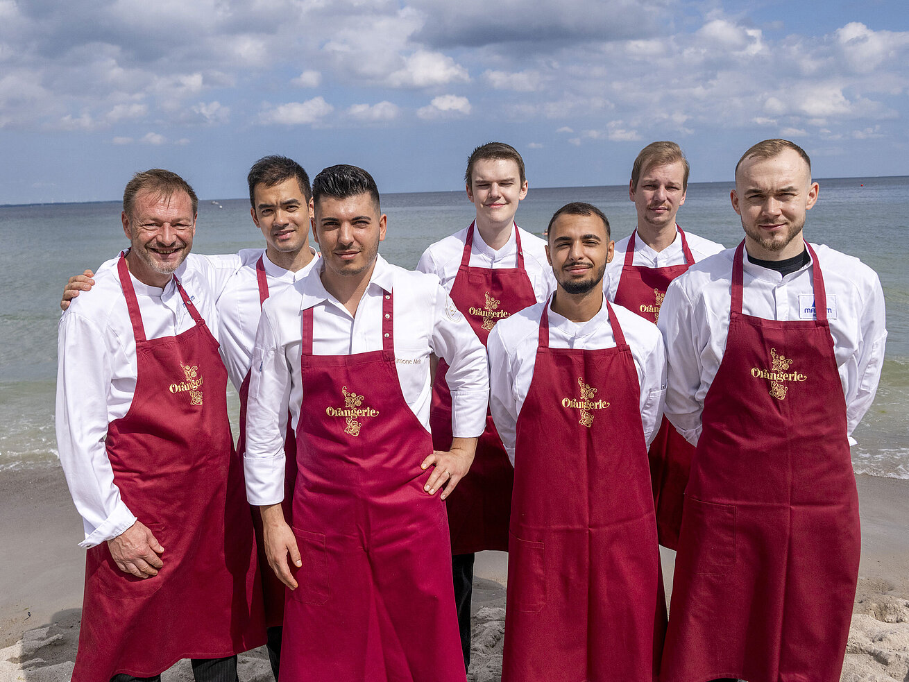 Küchenteam der Orangerie im Maritim Hotel Timmendorfer Strand, am Strand in Kochuniformen und roten Schürzen stehend.